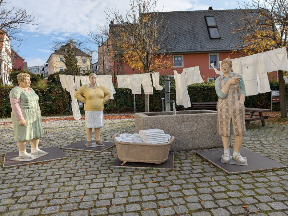 Skulpturen von drei Frauen beim Wäschewaschen im Hof mit aufgehängter Wäsche und Waschkasten